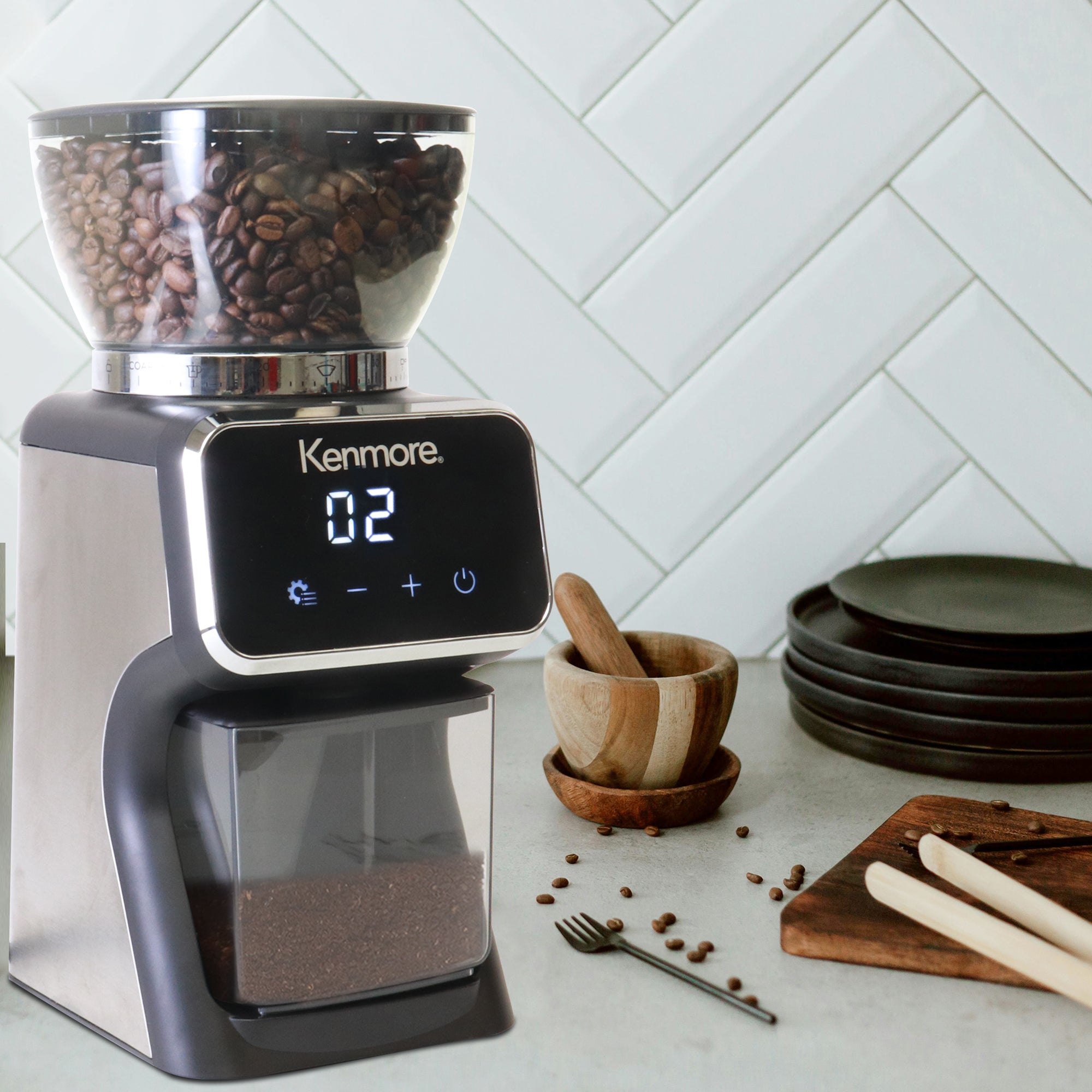 Kenmore stainless steel digital burr coffee grinder on a light gray marble countertop with a white tile backsplash behind and dishes arranged around it.