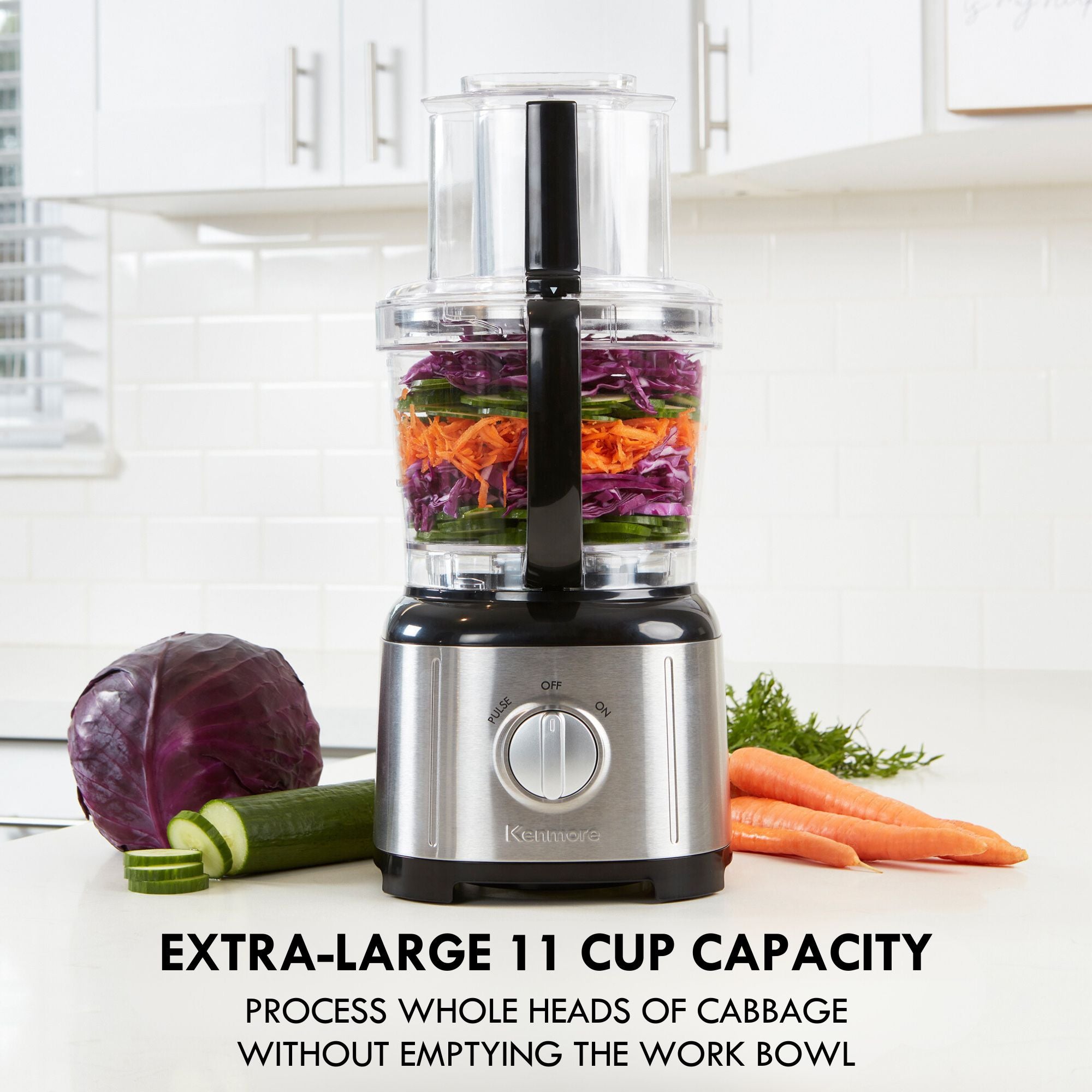 Kenmore 11 cup food processor and vegetable chopper on a light gray countertop with white tile backsplash and cupboards behind. There are chopped vegetables in the food processor and a whole red cabbage, partially sliced cucumber, carrots, and dill on either side. Text below reads, "Extra-large 11 cup capacity: Process whole heads of cabbage without emptying the work bowl"