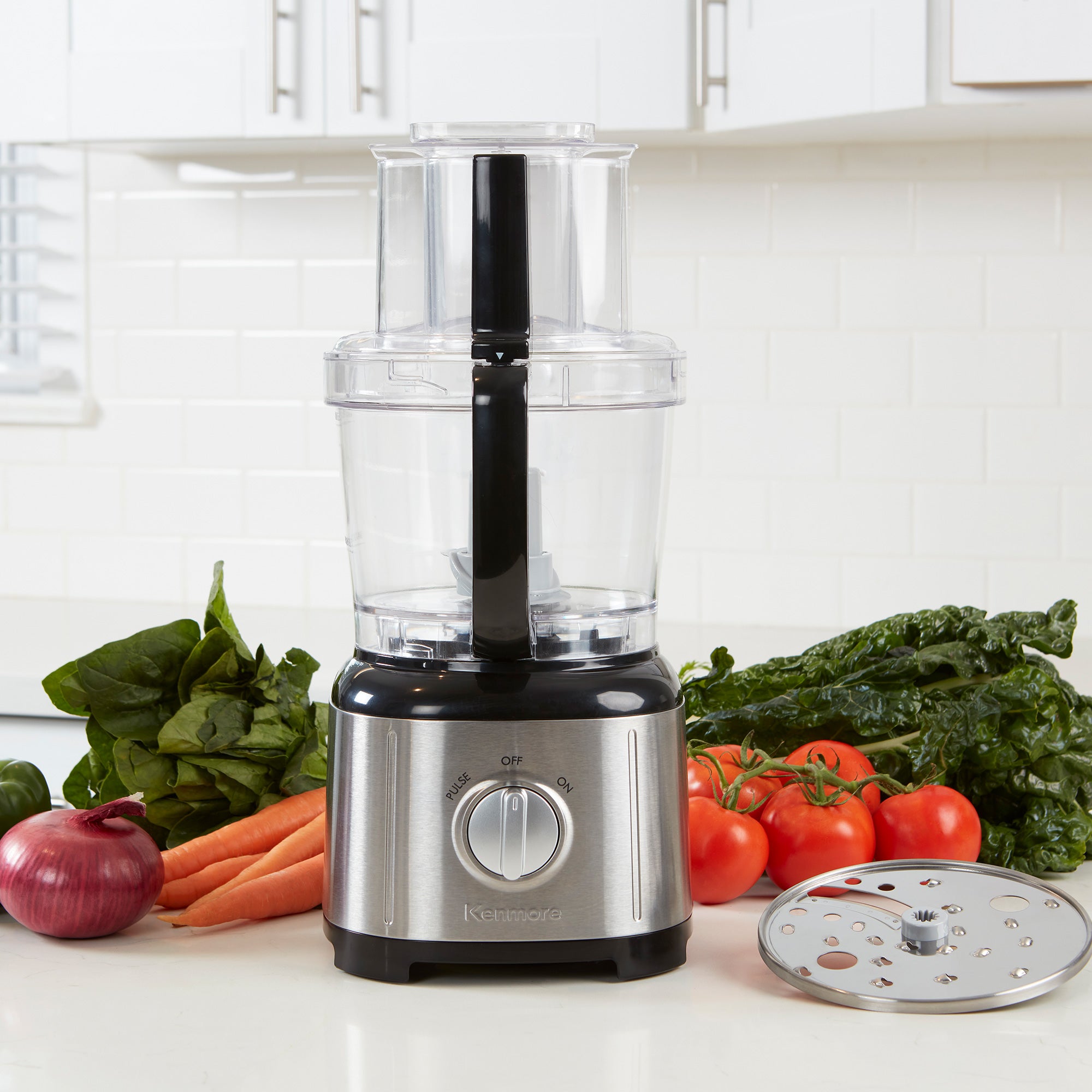 Kenmore 11 cup food processor and vegetable chopper, empty, on a light gray countertop with white tile backsplash behind. There is a bowl of chopped tomatoes to the left and a cutting board, knife, and whole and chopped ingredients to the right.