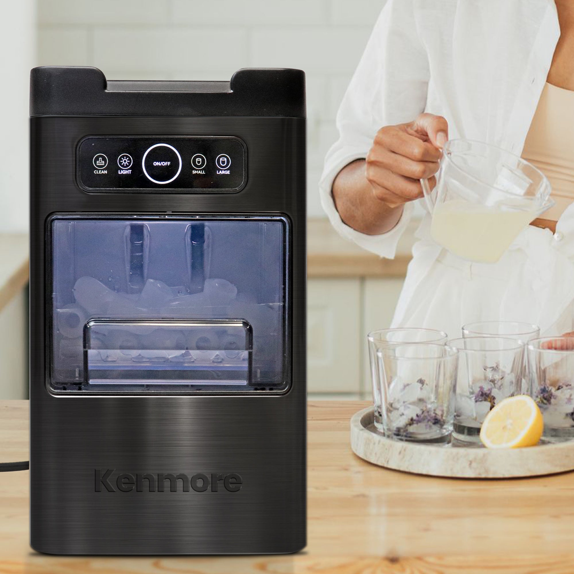 Kenmore automatic front-load ice maker on a light wooden countertop with a person pouring lemonade into glasses filled with ice in the background