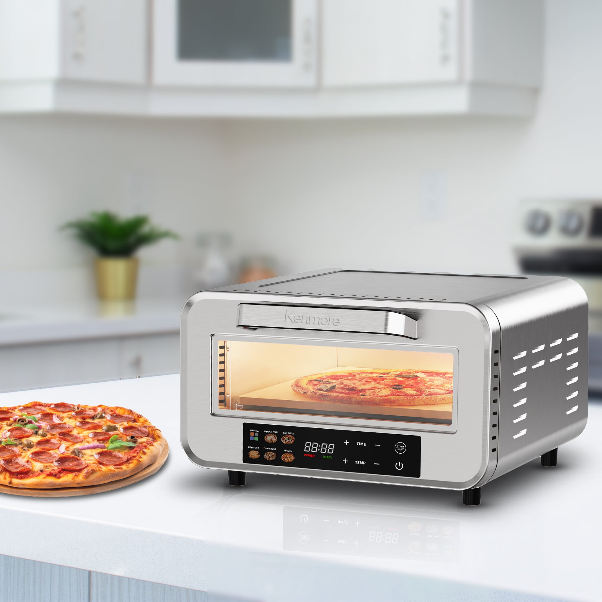 Kenmore portable countertop pizza oven with a pizza cooking inside and another beside it on a white kitchen island with white counter and cabinets in the background